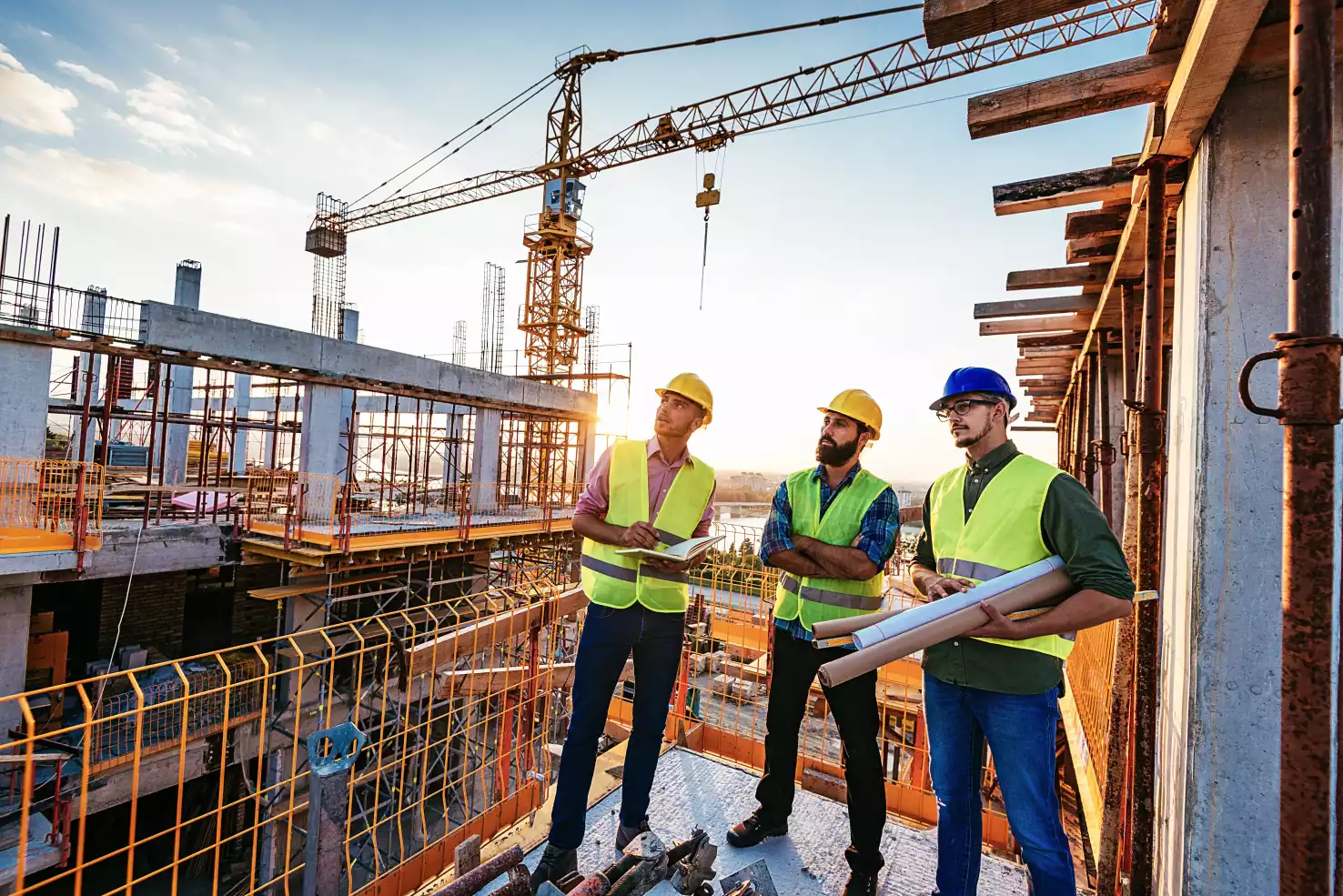Trois ingénieurs en gilets de sécurité discutent sur un chantier de construction au coucher du soleil, grue en arrière-plan.