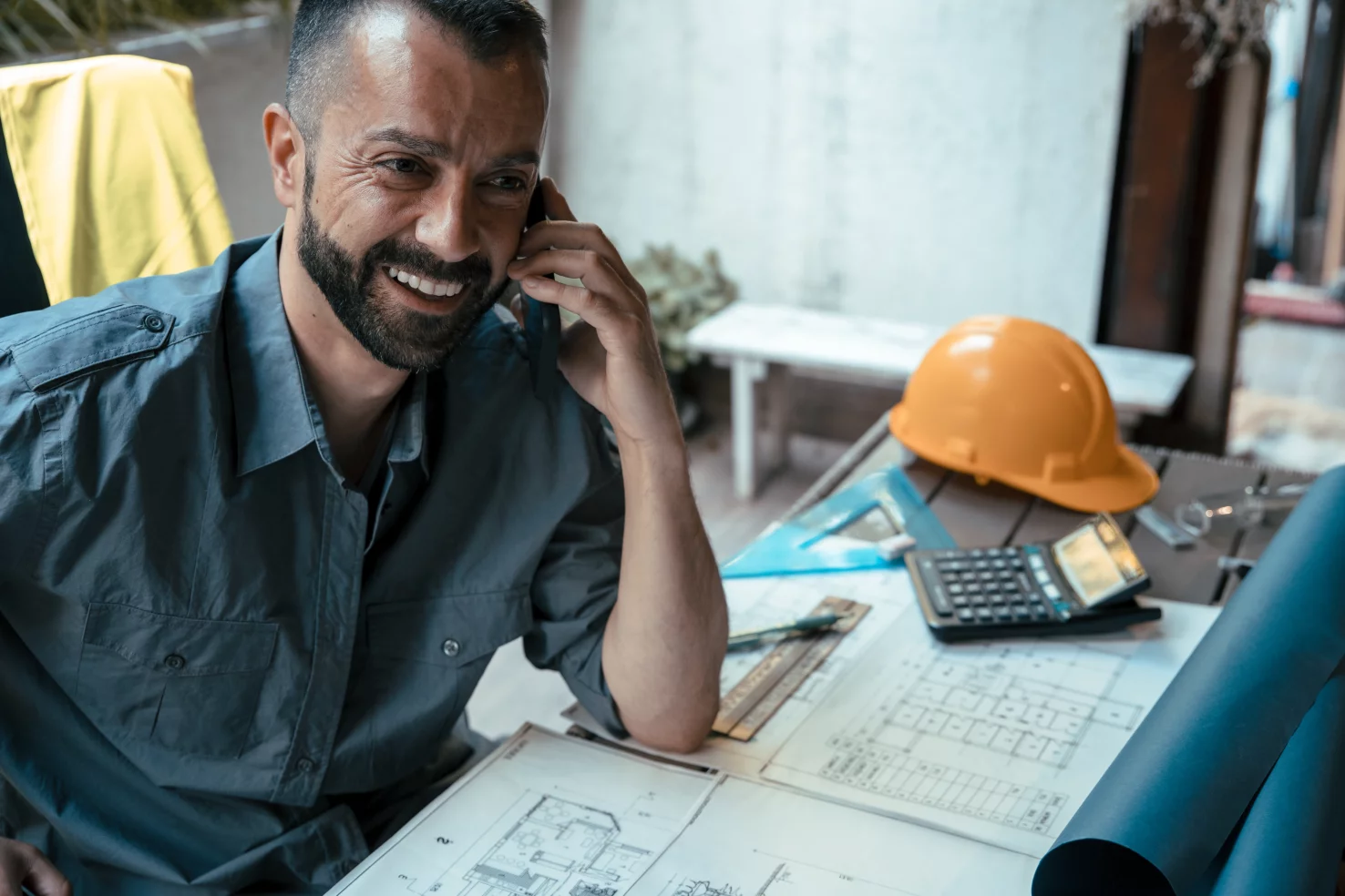 Architecte souriant au téléphone à son bureau, plans et casque de chantier devant lui.